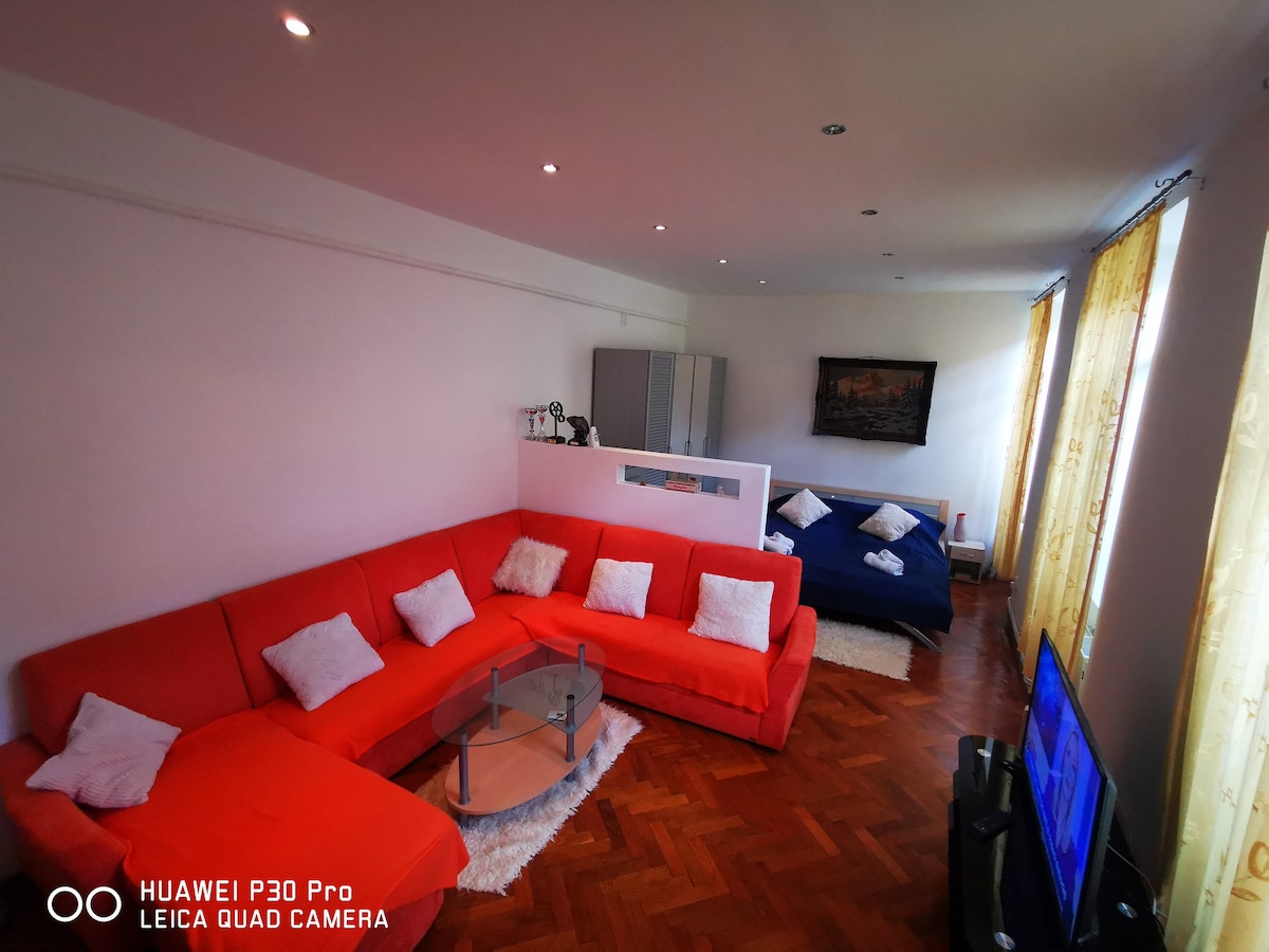 A spacious living area is furnished with a large red sectional sofa adorned with cushions. A glass coffee table is centrally placed on a soft area rug, while a flat-screen television is positioned nearby. Natural light filters through sheer curtains covering the windows.