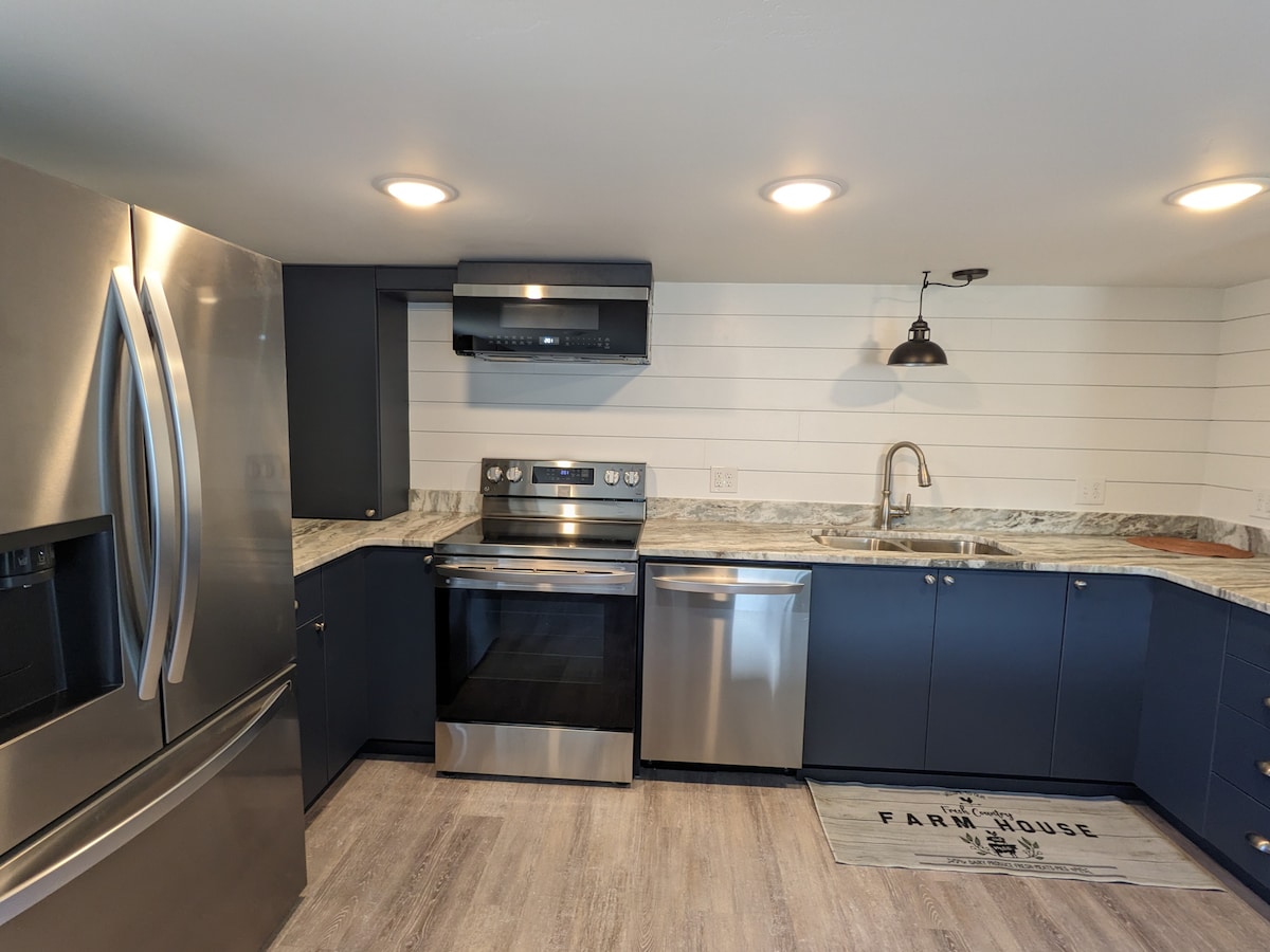 A modern kitchen features stainless steel appliances, including a refrigerator and an oven with a smooth cooktop. Dark cabinetry contrasts with the light marble countertops, while soft illumination from recessed lighting and a pendant lamp enhances the space. A farmhouse-style mat lies on the floor.
