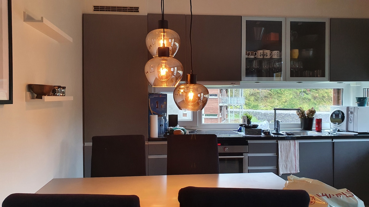 A modern kitchen features a dining table surrounded by dark chairs. Pendant lights hang above the table, illuminating the space. Cabinets and shelves, displaying various kitchenware, line the walls. A window offers natural light and a view of the outdoors.