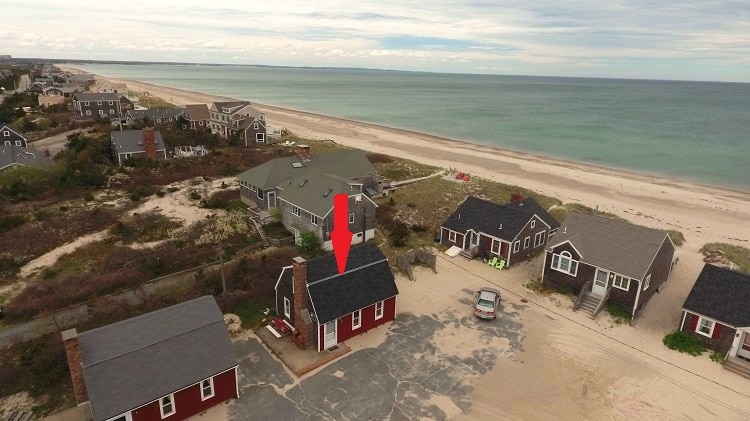 An aerial view captures the charming cottage, positioned near the beach, with the ocean extending in the background. The surrounding area features several cottages and sandy paths, showcasing a serene coastal environment. A few vehicles can also be seen parked nearby.