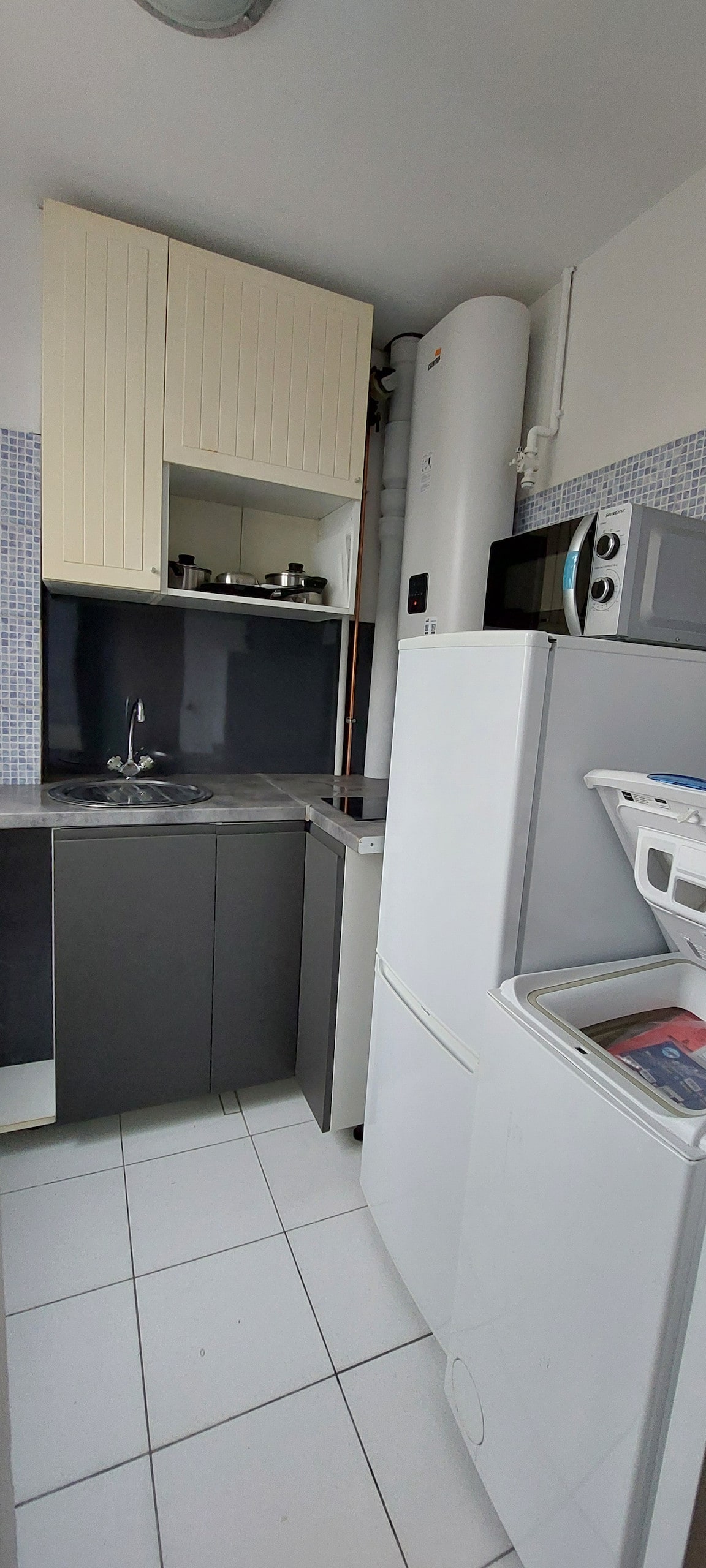 A compact kitchen space is displayed, featuring a combination of light wood and dark cabinetry. Essential appliances, including a refrigerator, stove, and sink, are arranged for functionality. The tiled floor adds a clean and modern touch, with small kitchenware visible on the shelves.