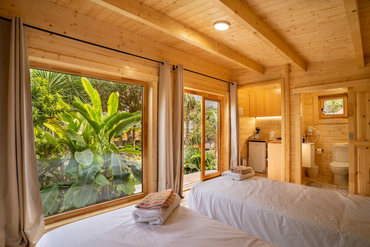A bright and airy sleeping area is framed by large windows overlooking a vibrant garden. Two beds are arranged beside the windows, with wooden beams enhancing the room’s natural aesthetic. A kitchenette and bathroom area are visible in the background.