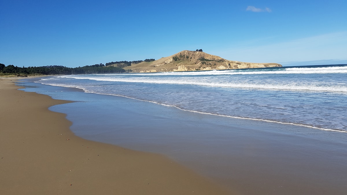 A serene coastal landscape displays a wide sandy beach meeting gentle waves under a clear blue sky. Rolling hills and greenery are visible in the background, enhancing the tranquil setting. The shoreline curves gracefully, inviting exploration and relaxation along the water's edge.
