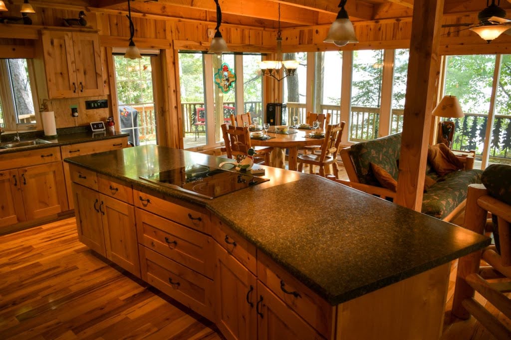 A spacious kitchen is presented, featuring wooden cabinetry and a large island with a dark countertop. Natural light floods the area through multiple windows, inviting views of the surrounding trees. A dining table and seats are arranged nearby, with a cozy seating area visible in the background.