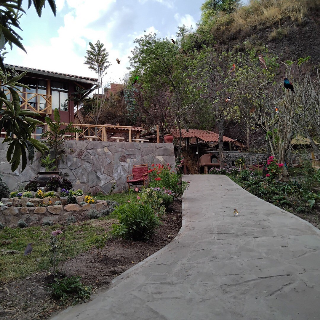 A winding pathway leads through a garden featuring colorful flowers and lush greenery. Natural stone walls frame the space, while a wooden structure is visible in the background, complemented by rustic seating areas. The atmosphere reflects a harmonious connection with the surrounding landscape.