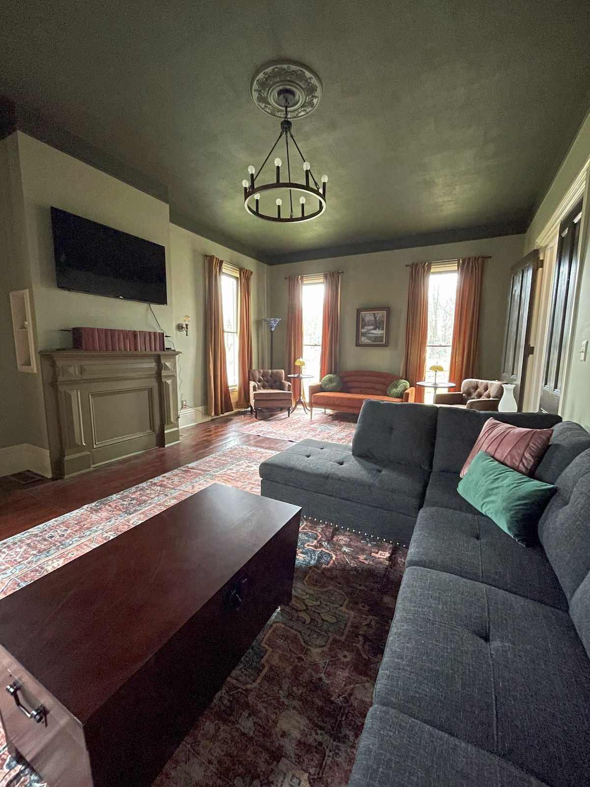 A comfortable seating area features a dark gray sectional sofa adorned with green and burgundy cushions. A wooden coffee table sits in the center, while large windows draped with rich curtains allow natural light to gently illuminate the space. A vintage-style chandelier hangs from the ceiling.