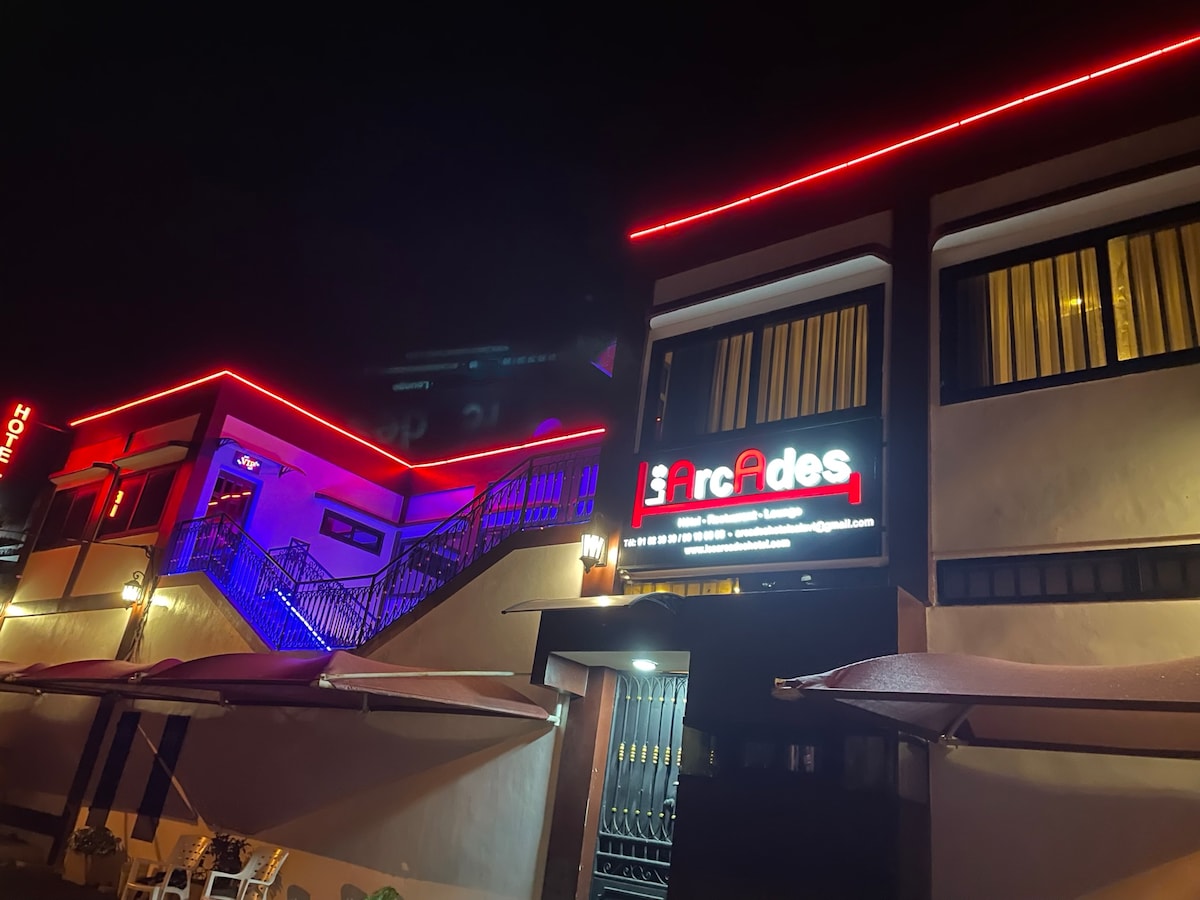 The exterior of the building is illuminated with colorful LED lighting, creating a vibrant evening atmosphere. Large windows line the second floor, revealing hints of the interior. A prominent sign displays the name of the establishment, while patio umbrellas are positioned outside.