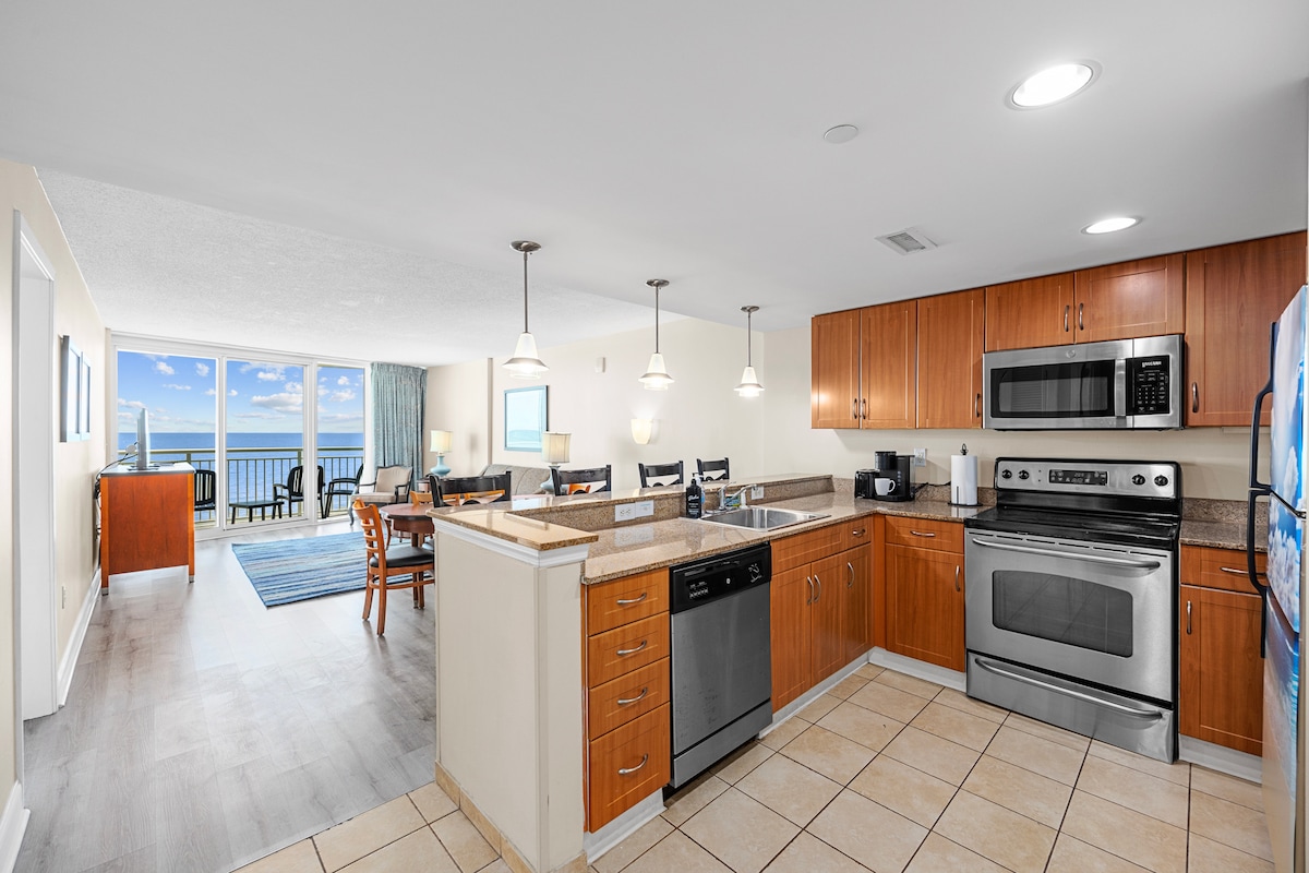 The image shows a modern kitchen with wooden cabinetry and stainless steel appliances, including a dishwasher, oven, and microwave. A granite countertop provides ample space, and a dining area with a table for four is visible in the background, framed by large windows offering ocean views.