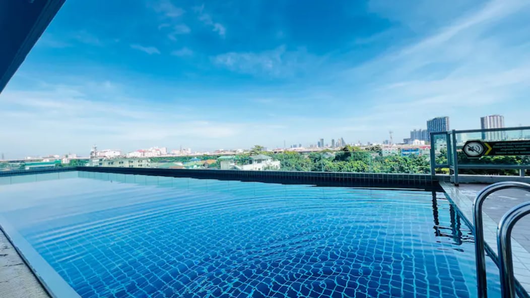 An inviting swimming pool reflects the clear blue sky, offering expansive views of the surrounding city landscape. The water's surface shimmers in the sunlight, inviting relaxation. A poolside railing and steps are visible, providing access to the water's edge.