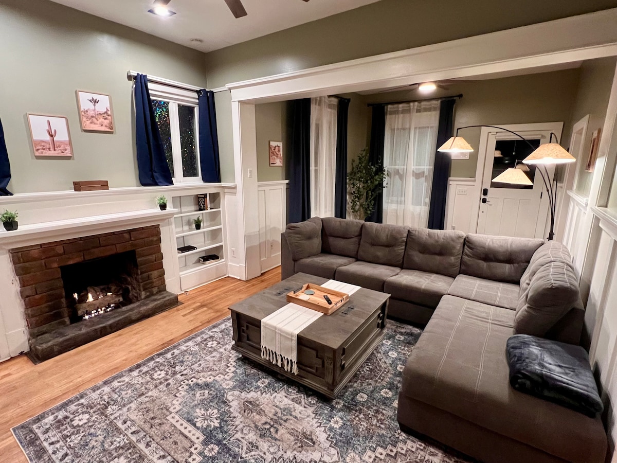 A spacious living area is highlighted by a sectional sofa, arranged around a coffee table with decorative items. A brick fireplace adds a cozy touch, while plants and artwork enhance the ambiance. Natural light enters through the windows, dressed with dark curtains.