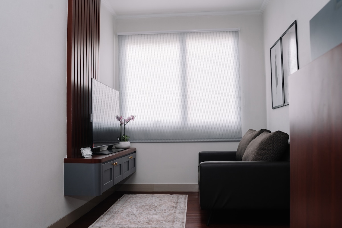 A cozy seating area features a dark sofa positioned against a light wall. A television is mounted on a stylish gray cabinet with a decorative plant displayed nearby. Natural light enters through a large window, complemented by simple curtains.