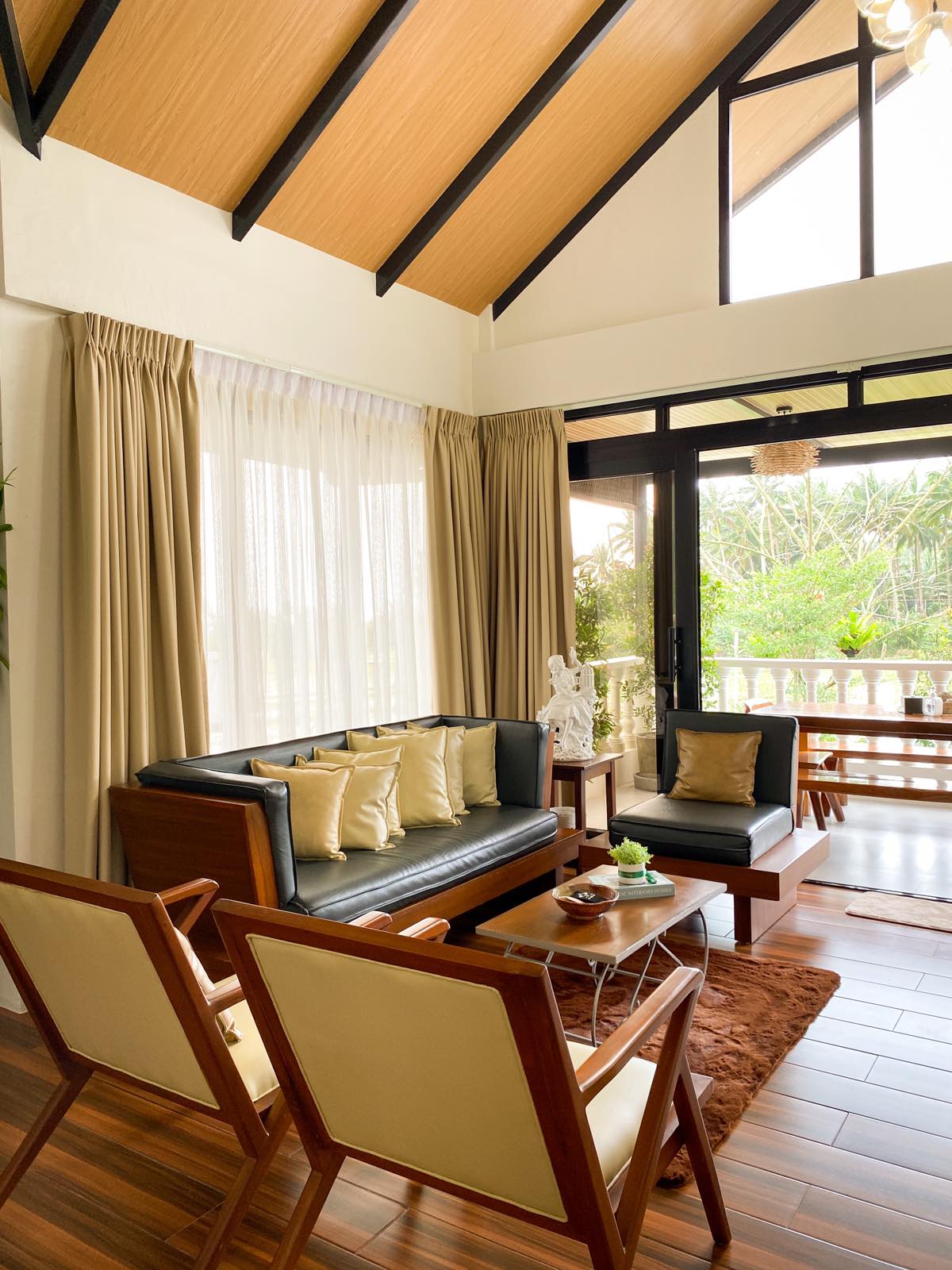 A stylish living area is presented with large windows allowing natural light to flood the space. Comfortable seating is arranged around a small table, complemented by decorative cushions. Exposed wooden beams enhance the ceiling's height, creating an open and airy atmosphere.