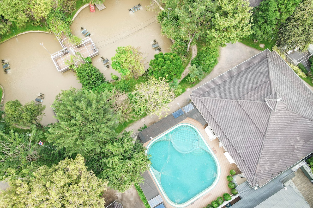 An aerial view captures a tranquil outdoor area featuring a swimming pool beside the house and a nearby pond. Surrounding greenery and lounge chairs enhance the natural setting, while pathways lead to various spots for relaxation amid lush foliage.