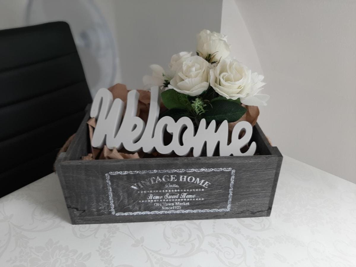 A rustic wooden box displays a decorative 'Welcome' sign alongside a small arrangement of artificial white roses. The box is filled with crinkled paper, enhancing the cozy presentation.