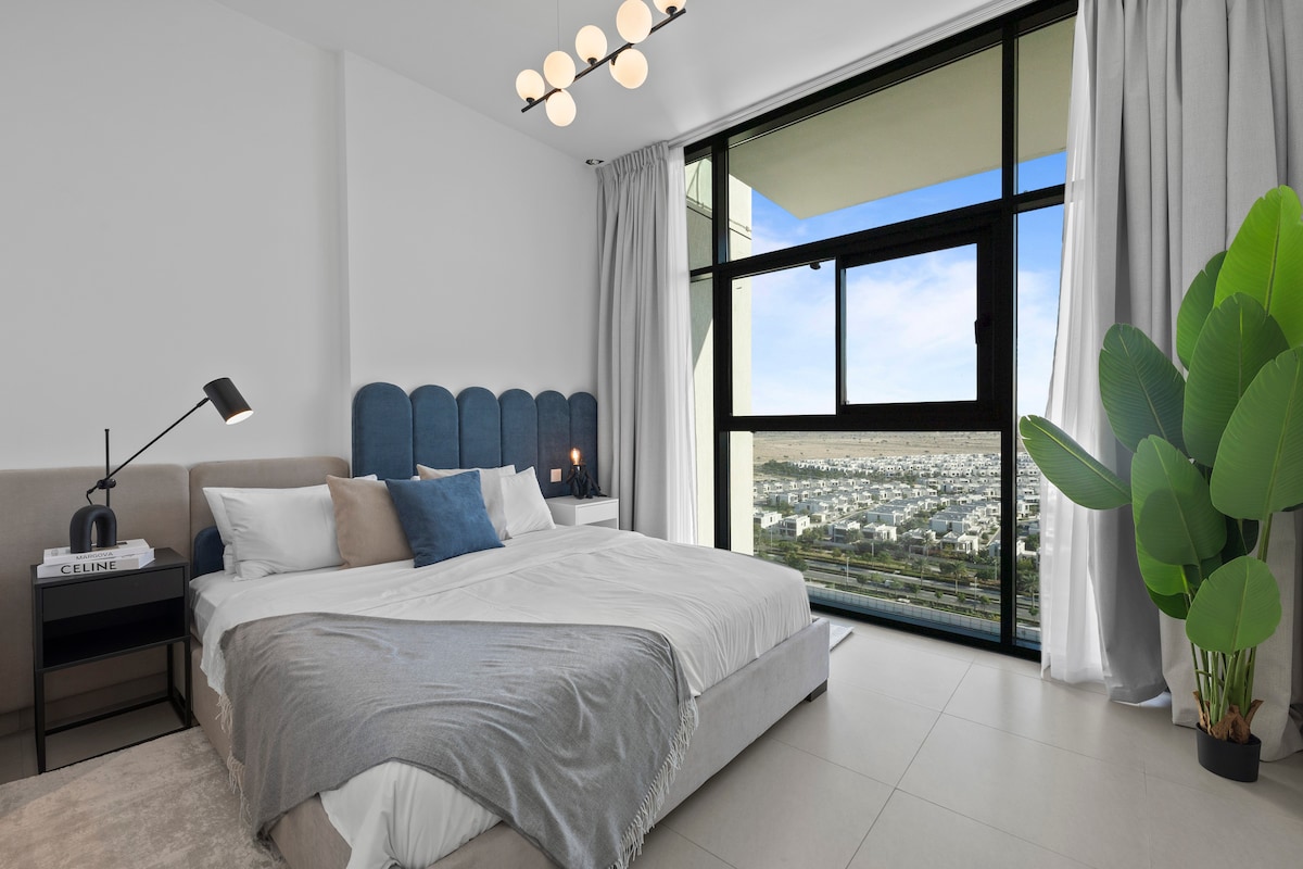 A spacious bedroom features a large bed with a soft grey blanket and decorative pillows, set against a blue upholstered headboard. Natural light pours in through a large window adorned with sheer curtains, offering expansive views of the cityscape. A bedside lamp and a decorative plant complete the ambiance.