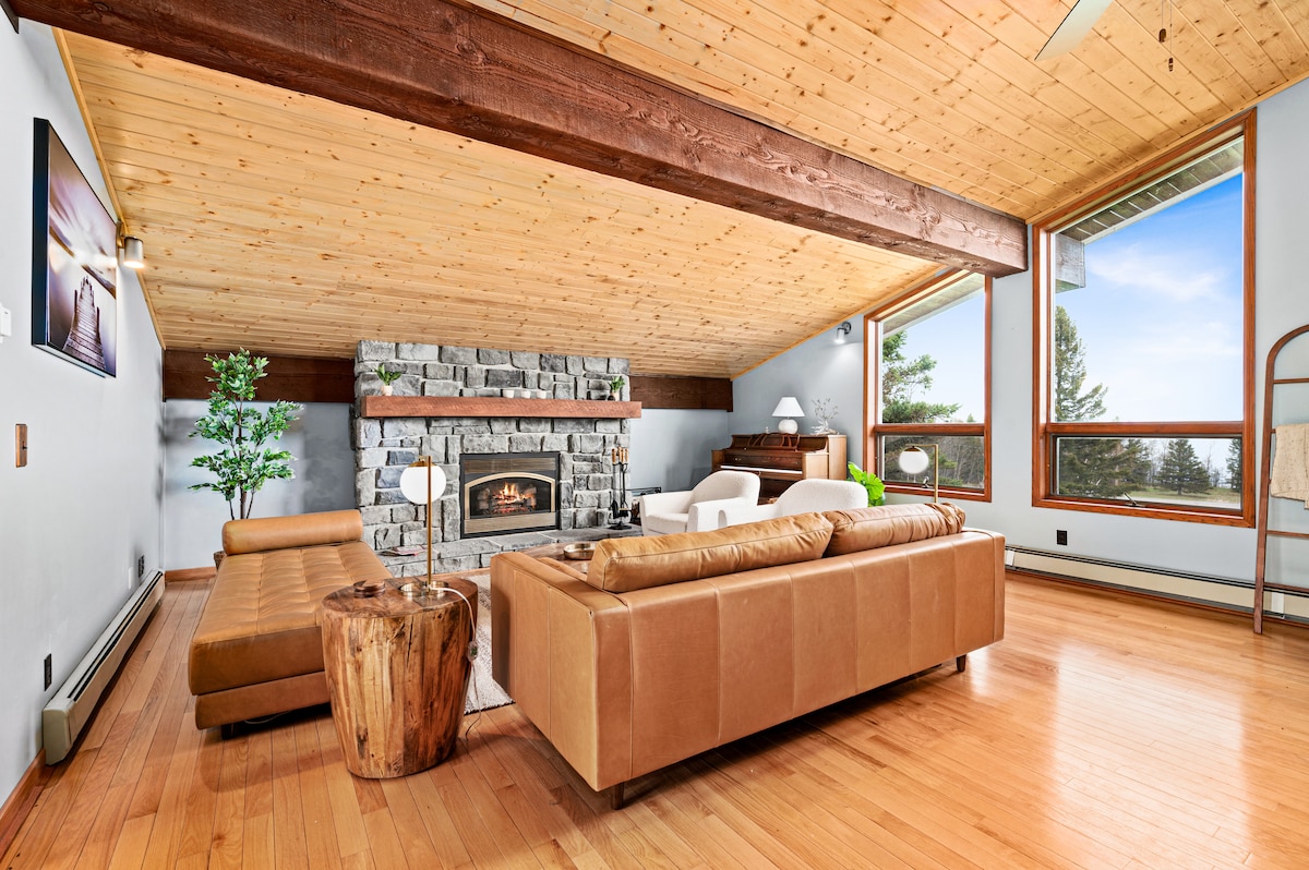 A warm-toned living area features a stone fireplace as its focal point, surrounded by large windows that provide views of the outdoors. Comfortable seating is arranged with a mix of leather and wooden furniture, while wooden beams extend across the ceiling, enhancing the spacious feel.