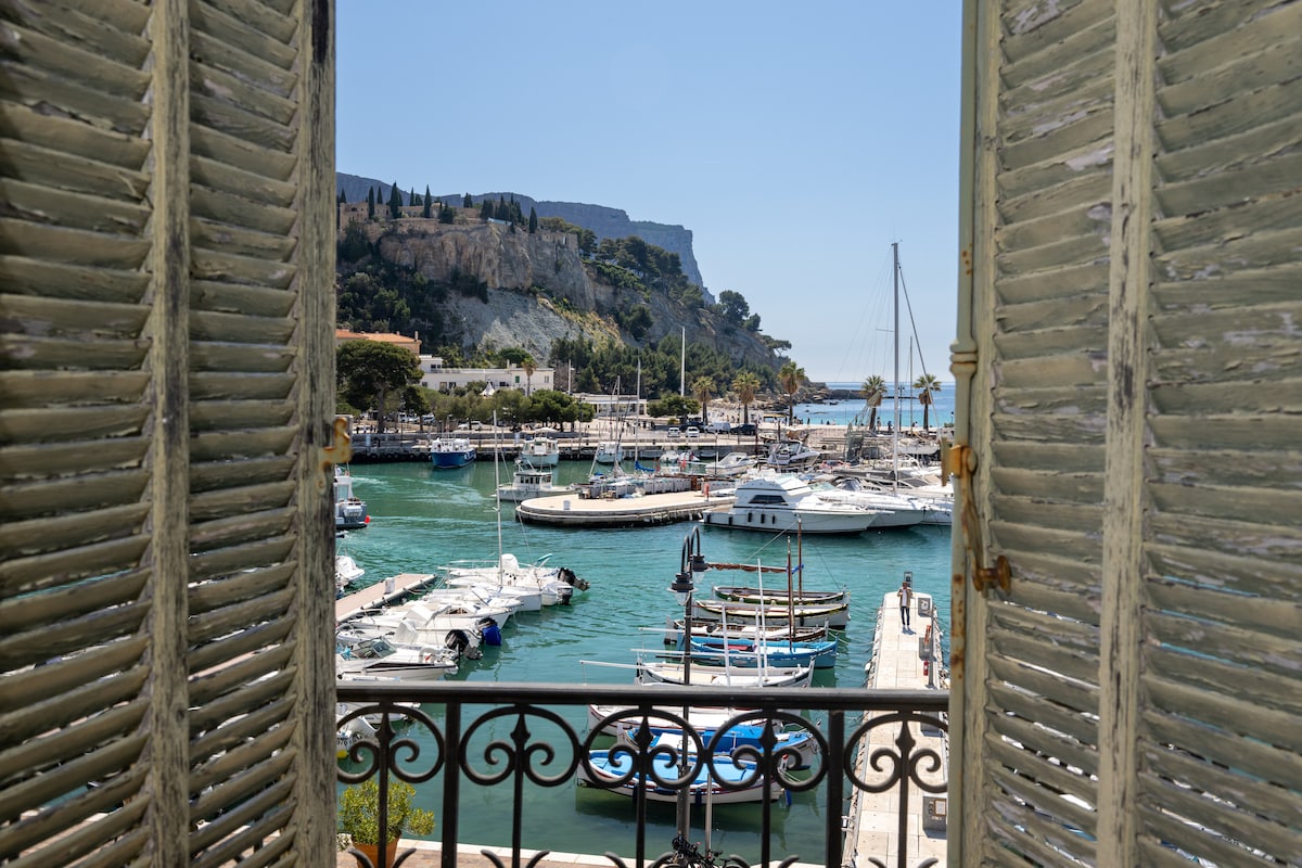 The view showcases a vibrant harbor filled with moored boats and yachts. Rolling hills and a distant cliff provide a natural backdrop, framed by open shutters that invite in natural light. The scene captures the essence of maritime life in a picturesque setting.