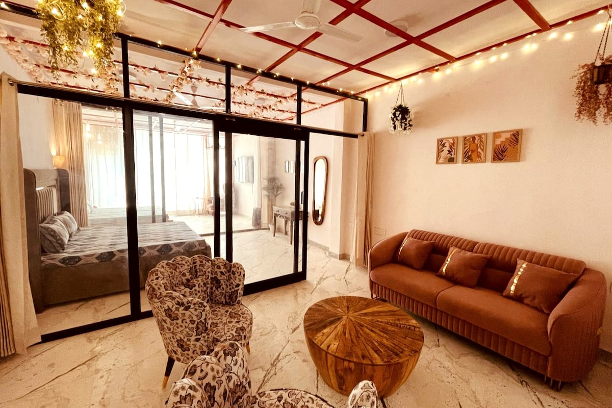 A bright living space is framed by glass sliding doors leading to a bedroom area. Soft seating is arranged around a circular wooden table, with patterned armchairs and a plush sofa. Ambient lighting is provided by string lights intertwined with greenery, adding warmth to the atmosphere.