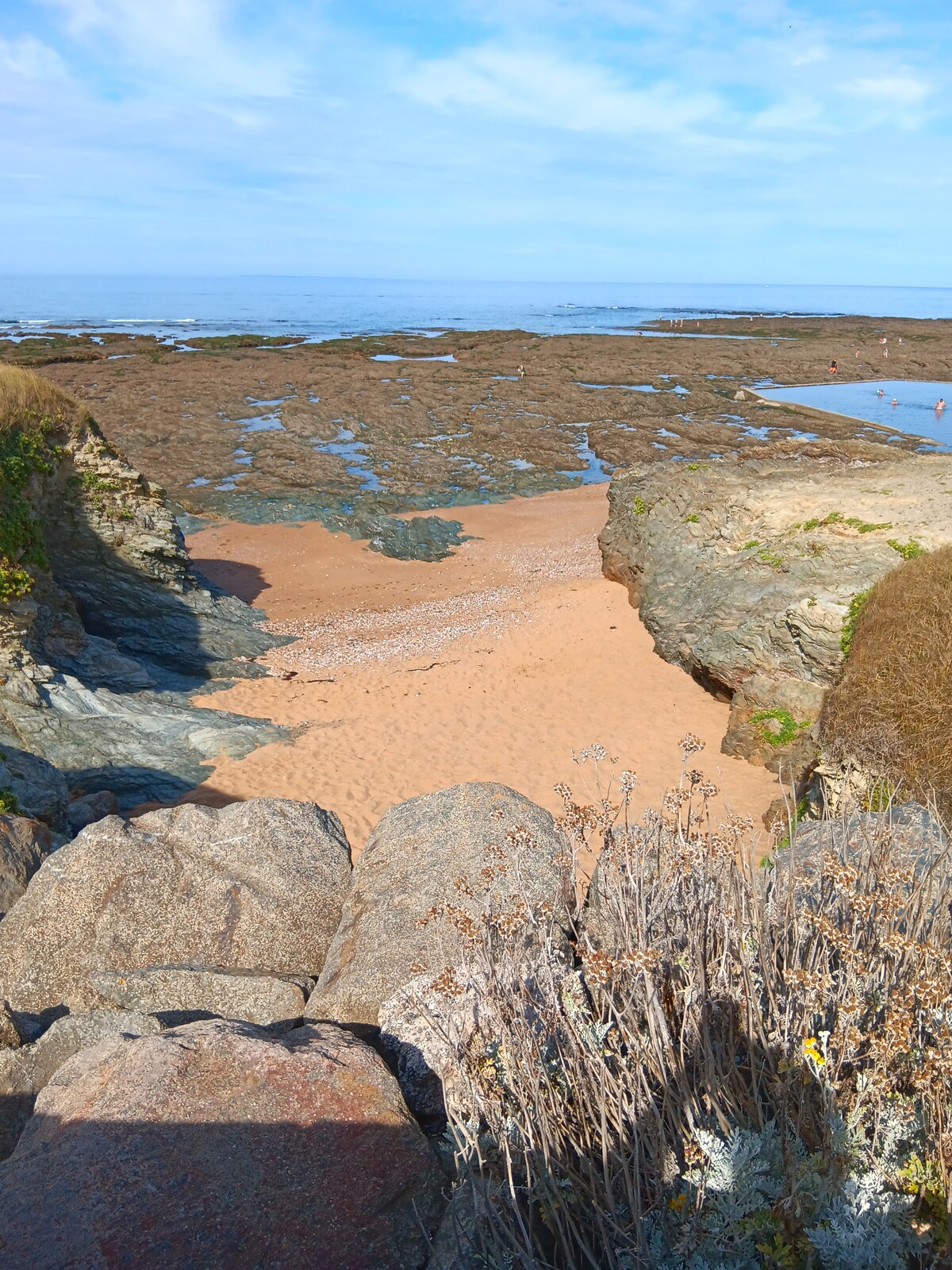 A sandy beach scene is framed by rocky outcrops and coastal vegetation. The tide is low, revealing stretches of rocky terrain and shallow waters. A clear sky overhead features scattered clouds, enhancing the coastal atmosphere. The beach is visible in the distance, inviting exploration.
