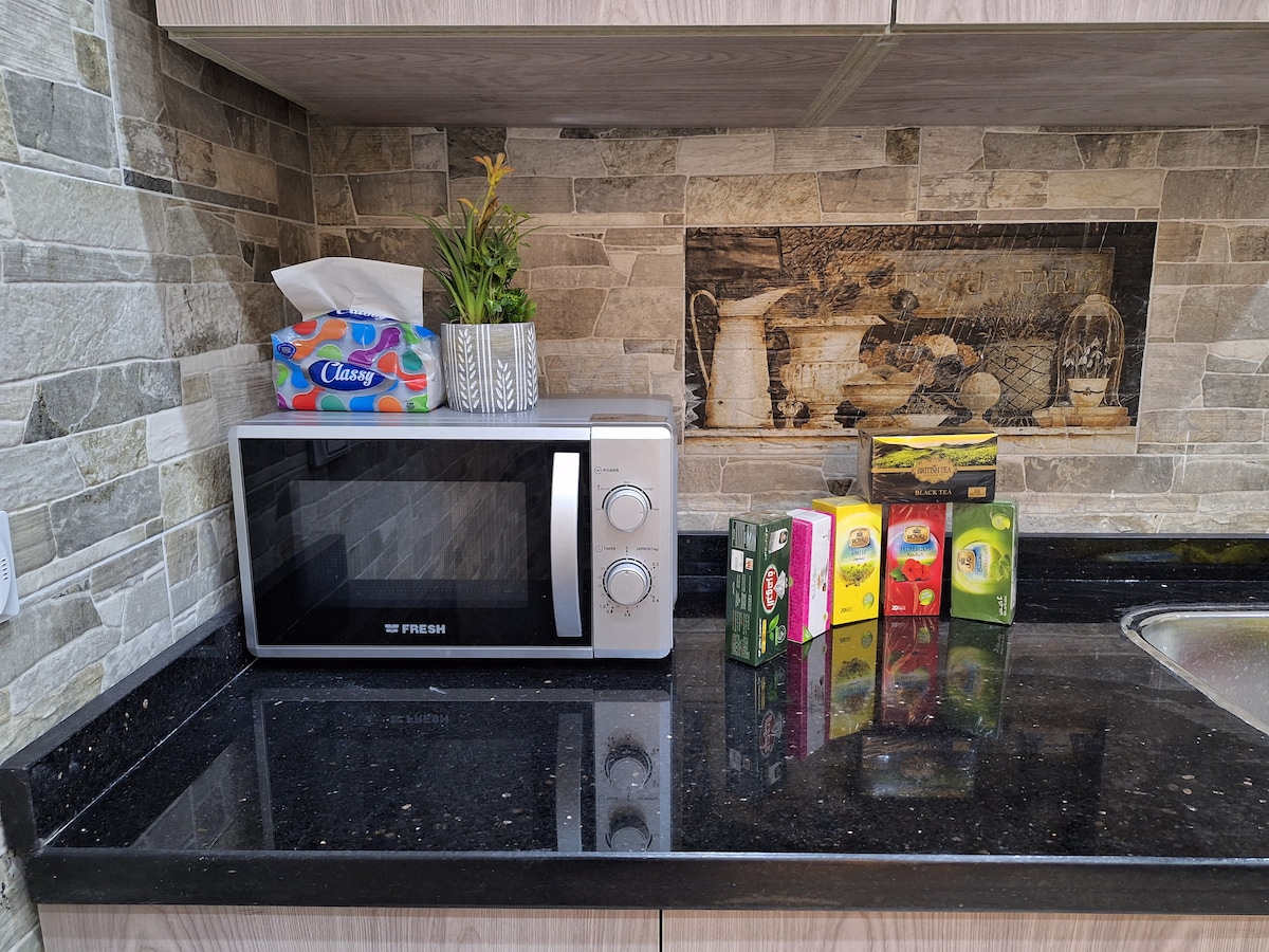 A modern kitchen countertop is shown featuring a microwave and a small potted plant. A box of tissues and several colorful tea boxes are arranged neatly beside the microwave, all resting on a polished black surface with decorative stone wall accents behind.