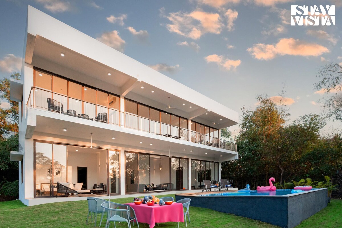 A modern villa is showcased, featuring large glass windows that provide garden views. An outdoor pool is visible alongside a vibrant seating area, complemented by a colorful tablecloth and dining setup. The spacious lawn and patio areas invite relaxation.