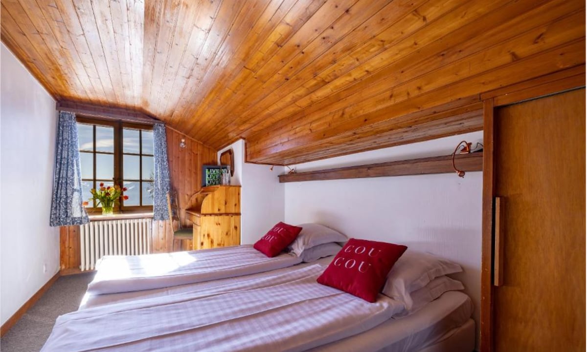 The room features a warm wooden ceiling and natural light entering through a large window adorned with floral curtains. Two spacious beds are set apart, each topped with red cushions. A wooden dresser and a closet add to the room's functionality.