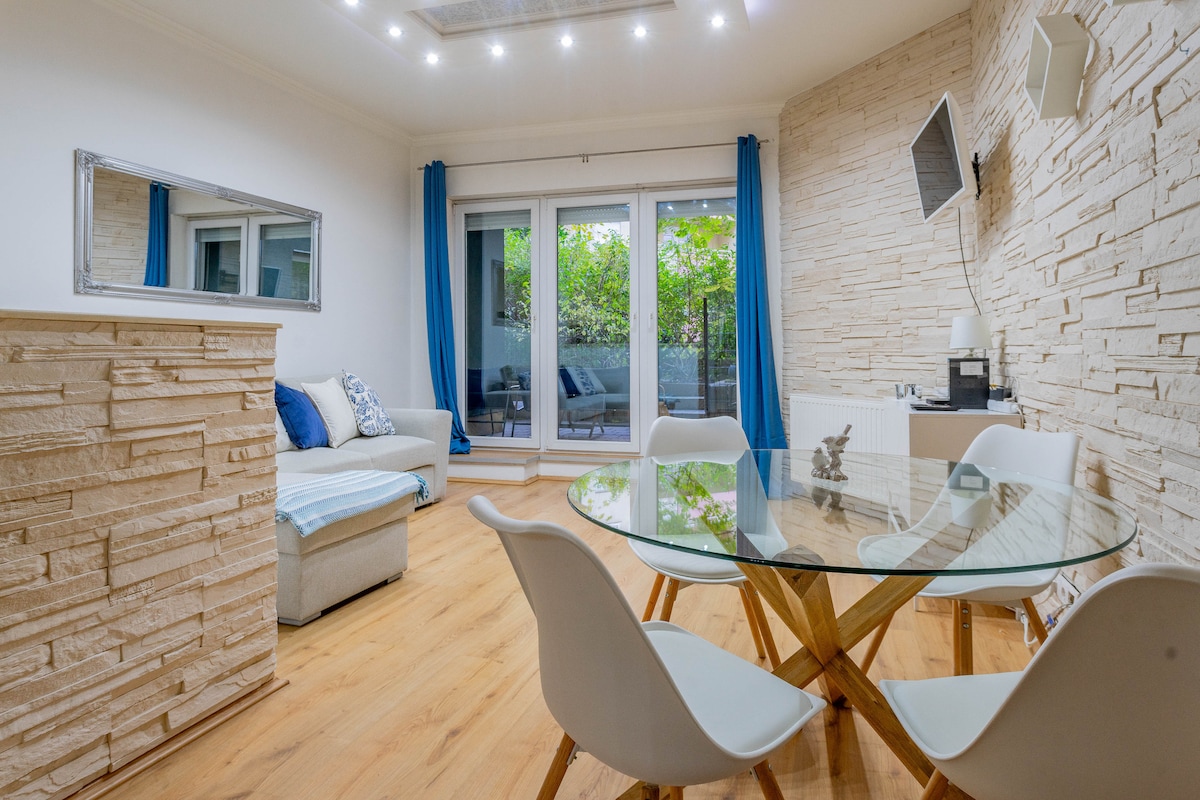 A modern living area features a glass dining table surrounded by four white chairs. A soft sofa is positioned nearby, and large sliding glass doors lead to a private outdoor space, framed by blue curtains. Natural light is enhanced by recessed ceiling lights.