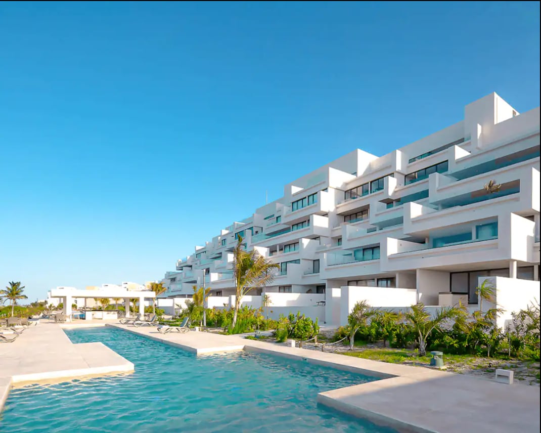 A modern multi-story building features a sleek design with spacious balconies. A long swimming pool runs alongside the structure, surrounded by lush greenery. Sun loungers line the pool area, creating an inviting space for relaxation. Clear blue skies enhance the property’s contemporary aesthetic.