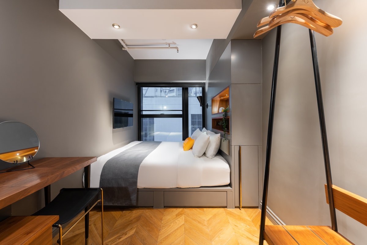 The modern room features a spacious bed with high-quality bedding, complemented by a sleek grey headboard. A dedicated workspace with a desk and chair is positioned nearby. Natural light filters through large windows, illuminating stylish furniture and wooden flooring.