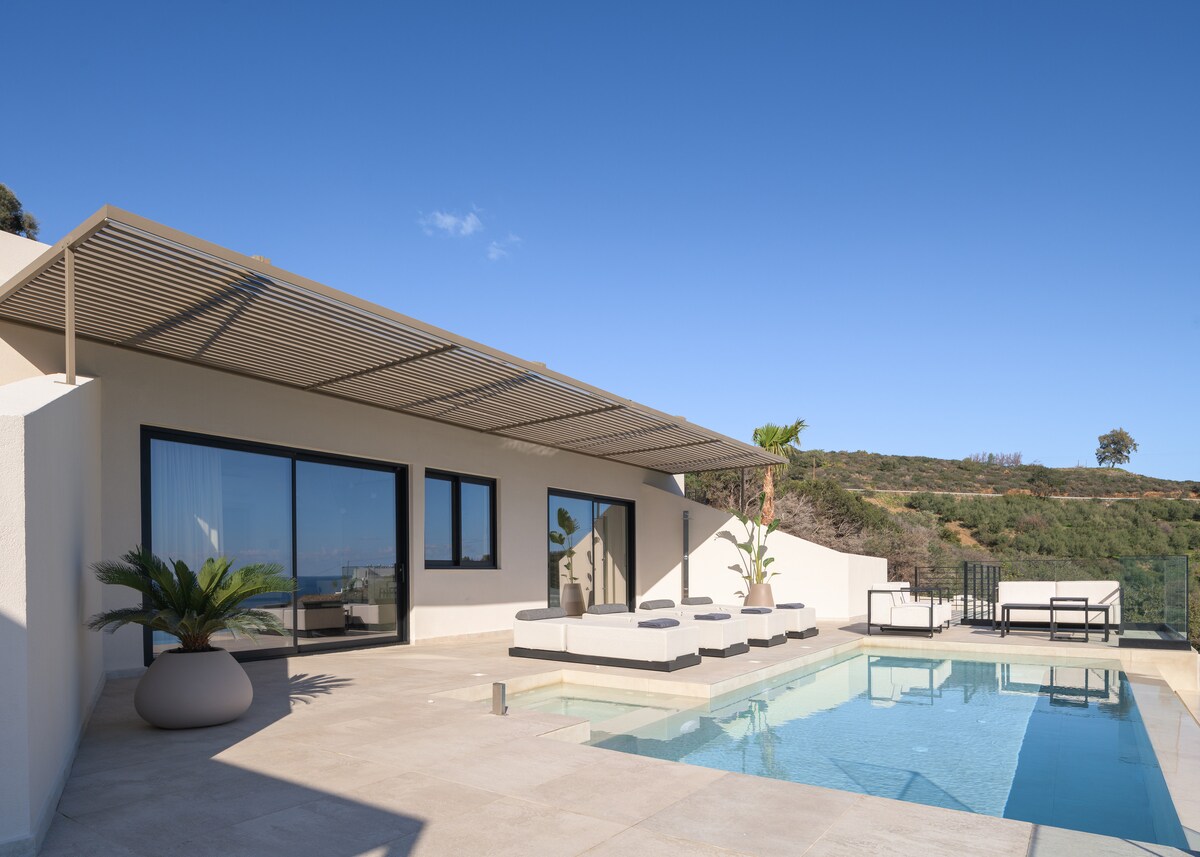 An outdoor area features a private infinity pool surrounded by a spacious terrace, complemented by modern sun loungers. The villa's large windows provide views of the surrounding landscape. A shaded seating area enhances the available outdoor space, inviting relaxation and enjoyment.