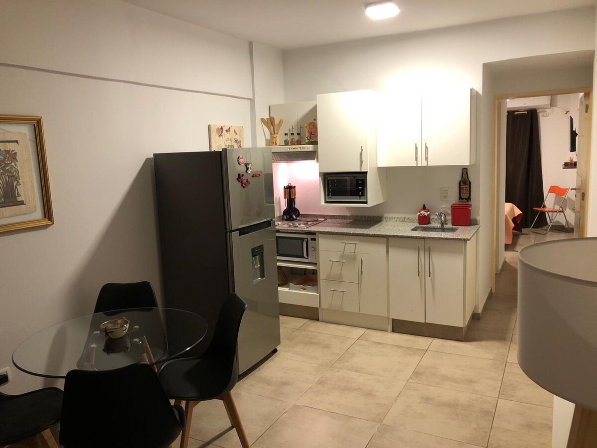 A modern kitchen area is depicted, featuring a stainless steel refrigerator and an oven with a glass cooktop. Light-colored cabinets support a countertop with a coffee maker and a blender. A round glass dining table with black chairs is positioned nearby, with a doorway leading to a separate room.
