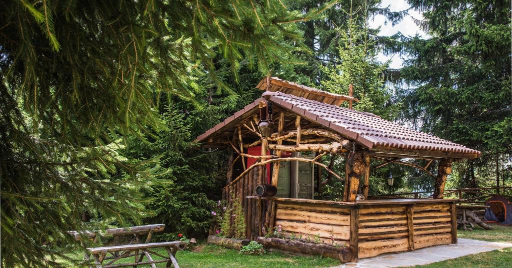 A rustic cabin made from natural wood is framed by tall, green trees. The structure features a sloped roof and an open terrace with wooden railings. Nearby, a simple seating area is comprised of wooden benches, surrounded by lush grass.