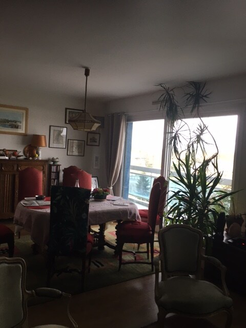 A dining area is set with a round table surrounded by upholstered chairs. Large windows allow natural light to fill the space, complemented by an assortment of indoor plants. Decorative wall art and a vintage sideboard add character to the room.