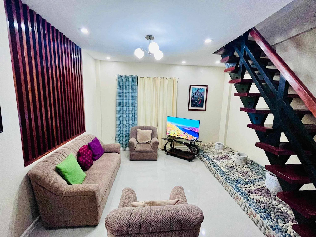 A well-lit living area features a comfortable sofa with colorful cushions and a pair of armchairs. A television is positioned on a low table, with a decorative stone arrangement along the wall. Natural light filters through the curtains, enhancing the warm atmosphere.