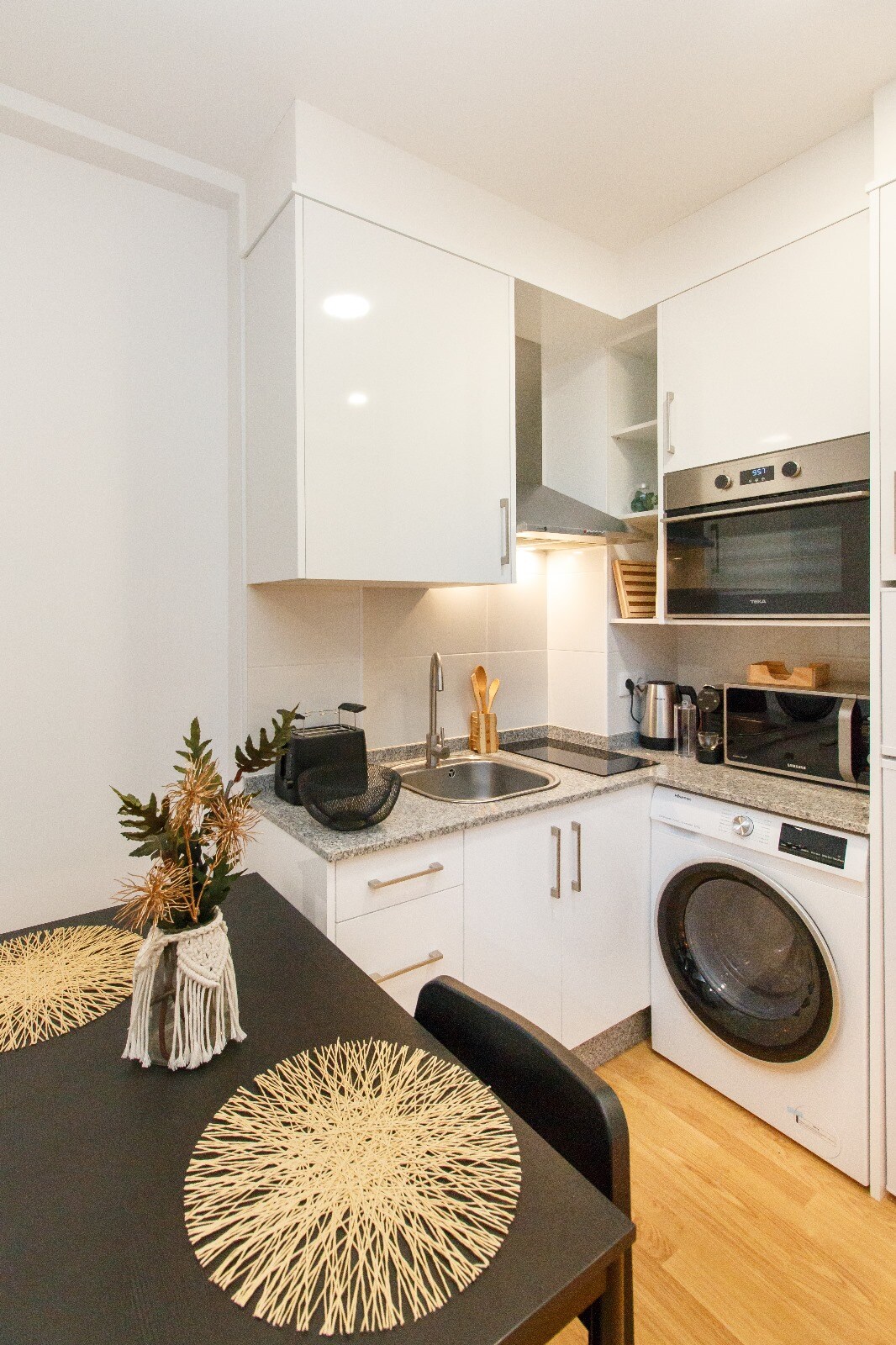 A modern kitchen is equipped with essential appliances, including a washing machine and a microwave. A small dining table with decorative placemats is positioned beside the kitchen area, enhancing the functional space. Sleek white cabinetry and granite countertops provide a clean and organized appearance.