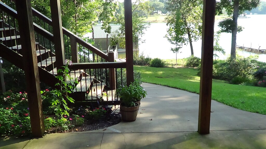 A serene outdoor space is presented, featuring a smooth concrete patio beneath wooden pillars. Lush greenery surrounds the area, with flowering plants in pots and well-maintained grass. A staircase leads to a higher deck, providing views of Lake Hartwell in the background.