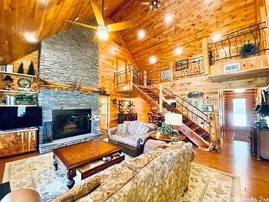 A spacious living area features a high, vaulted wooden ceiling with a ceiling fan. A large stone fireplace serves as a focal point, surrounded by rustic decor. A comfortable couch and wooden coffee table are present, alongside a staircase leading to the upper level.