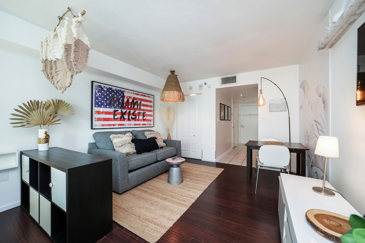 A modern living area is showcased with a gray sofa against a wall featuring an artistic American flag mural. A light-colored area rug is positioned on the dark wood floor, complemented by decorative lighting and a small dining table in the corner.