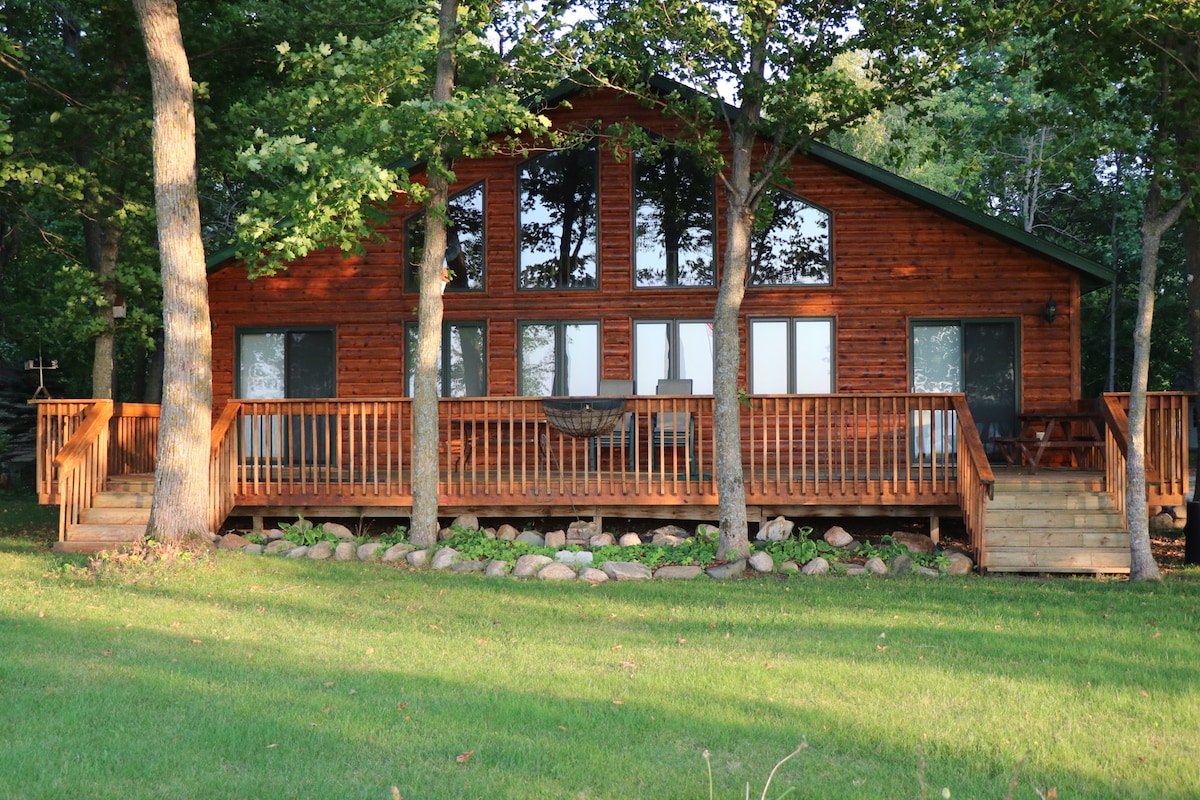 A spacious cabin features a wooden exterior, complemented by large windows that provide ample natural light. The front porch, surrounded by greenery, includes a railing and steps leading to the yard. Several trees are positioned closely, enhancing the outdoor setting.