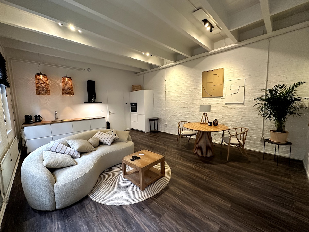 An open living space features a modern, curved sofa paired with decorative cushions. A round dining table surrounded by wooden chairs sits nearby. A plant adds a touch of greenery, while a kitchen area with sleek cabinetry is visible in the background.