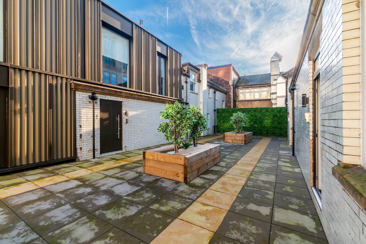 An outdoor courtyard is presented, featuring a mix of paving stones and wooden planters. Lush greenery is visible, with a backdrop of modern and traditional architecture. Bright skies enhance the open atmosphere of this inviting space.