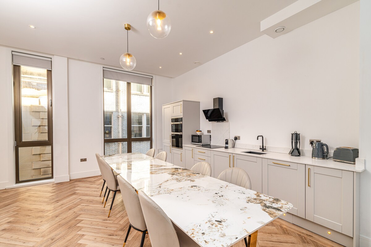 A modern kitchen area is highlighted by a large marble dining table surrounded by six upholstered chairs. Grey cabinetry lines one wall, equipped with built-in appliances. Natural light enters through large windows, reflecting off the sleek surfaces.