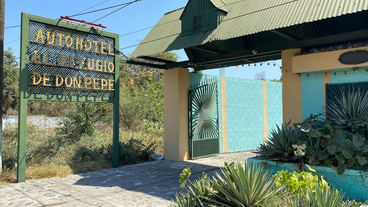 The entrance of the eco-friendly hotel is marked by a large sign detailing its name in bold lettering. The gate is flanked by vibrant greenery and the structure features a distinctive green roof, adding a unique character to the welcoming exterior.