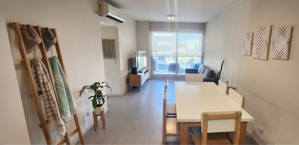 A bright and spacious living area is visible, featuring a dining table for six and a comfortable sofa. A TV unit rests against the wall, while large windows allow natural light to fill the room. A plant and wall decorations enhance the inviting atmosphere.