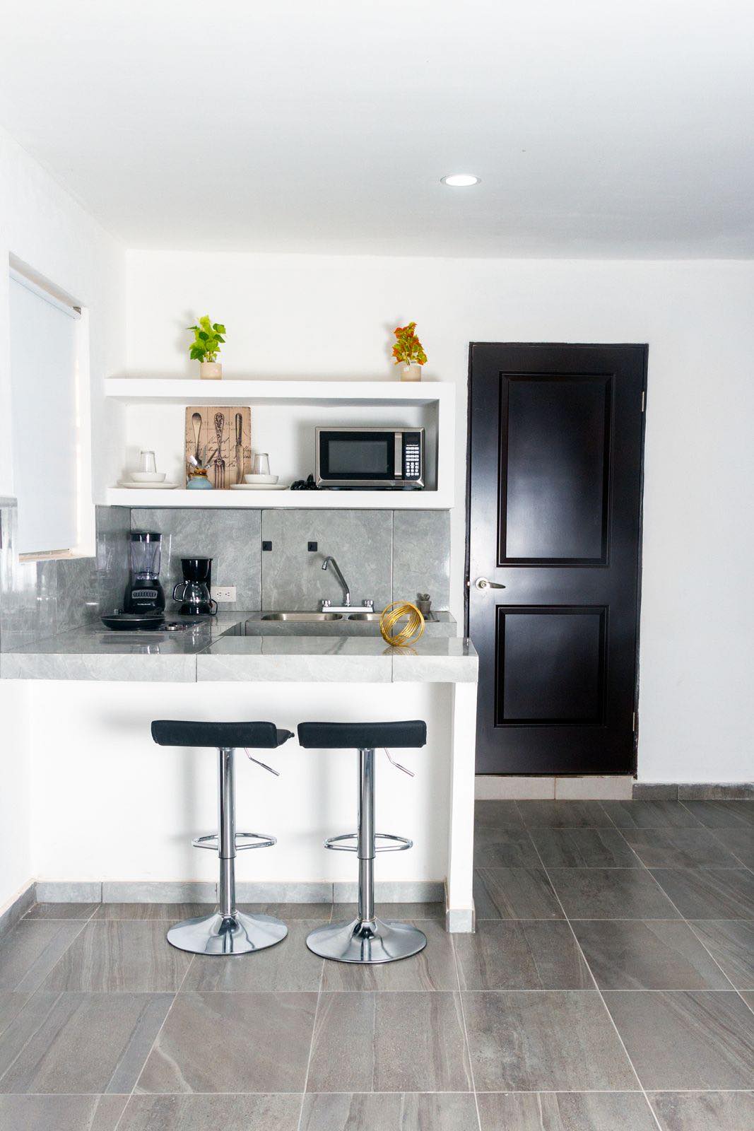 A modern kitchenette is equipped with a sink and microwave, complemented by a coffee maker and kitchen utensils displayed neatly. Two metal bar stools are positioned at the granite counter, contributing to the space's functionality and style. The door leads to another room, enhancing privacy.