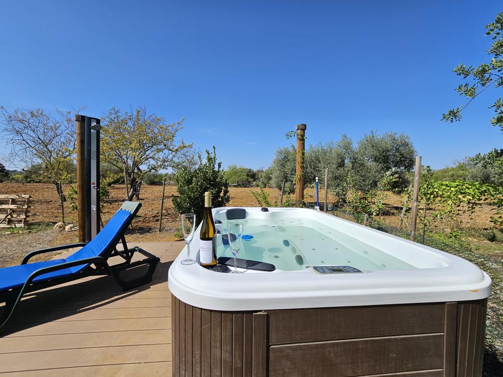 An outdoor jacuzzi is positioned on a wooden deck, surrounded by verdant fields and trees. A blue lounge chair sits nearby. Two glasses and a bottle of wine are placed on the edge of the jacuzzi, inviting relaxation amidst nature's tranquility.