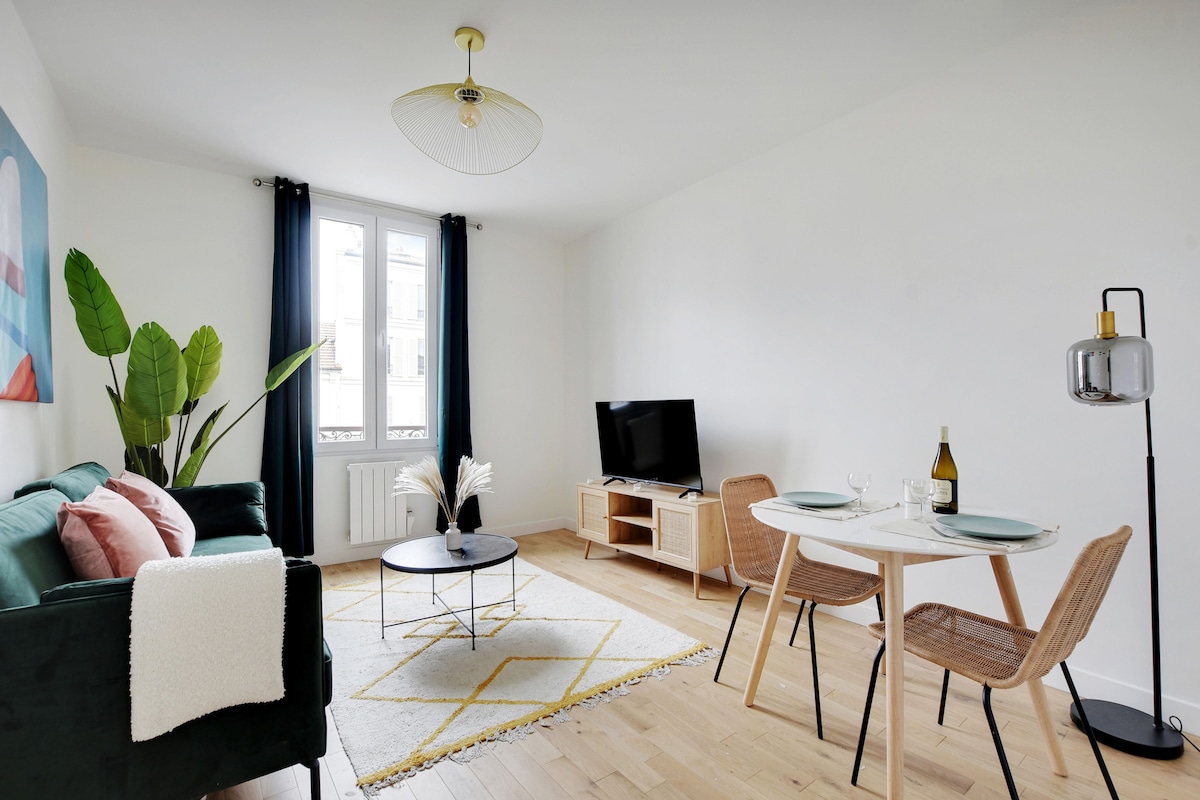 A light-filled living area features a green sofa accompanied by decorative pillows. A small round coffee table is positioned on a patterned rug. A dining table for two is nearby, set with dinnerware and a bottle of wine. Large windows are adorned with dark curtains.