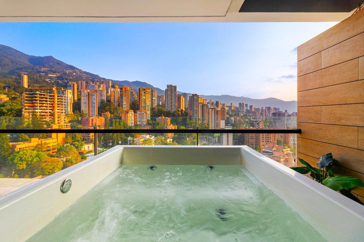 A private jacuzzi is situated on a wraparound terrace, offering views of the Medellin skyline and surrounding mountains. The clear water reflects the soft hues of the sunset, while the backdrop features tall buildings that outline the cityscape.