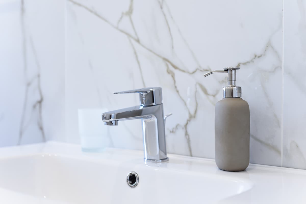 A modern bathroom sink area is shown, featuring a sleek chrome faucet alongside a minimalist grey soap dispenser. The countertop is smooth and clean, complemented by marble-patterned wall tiles and a softly glowing glass tumbler.