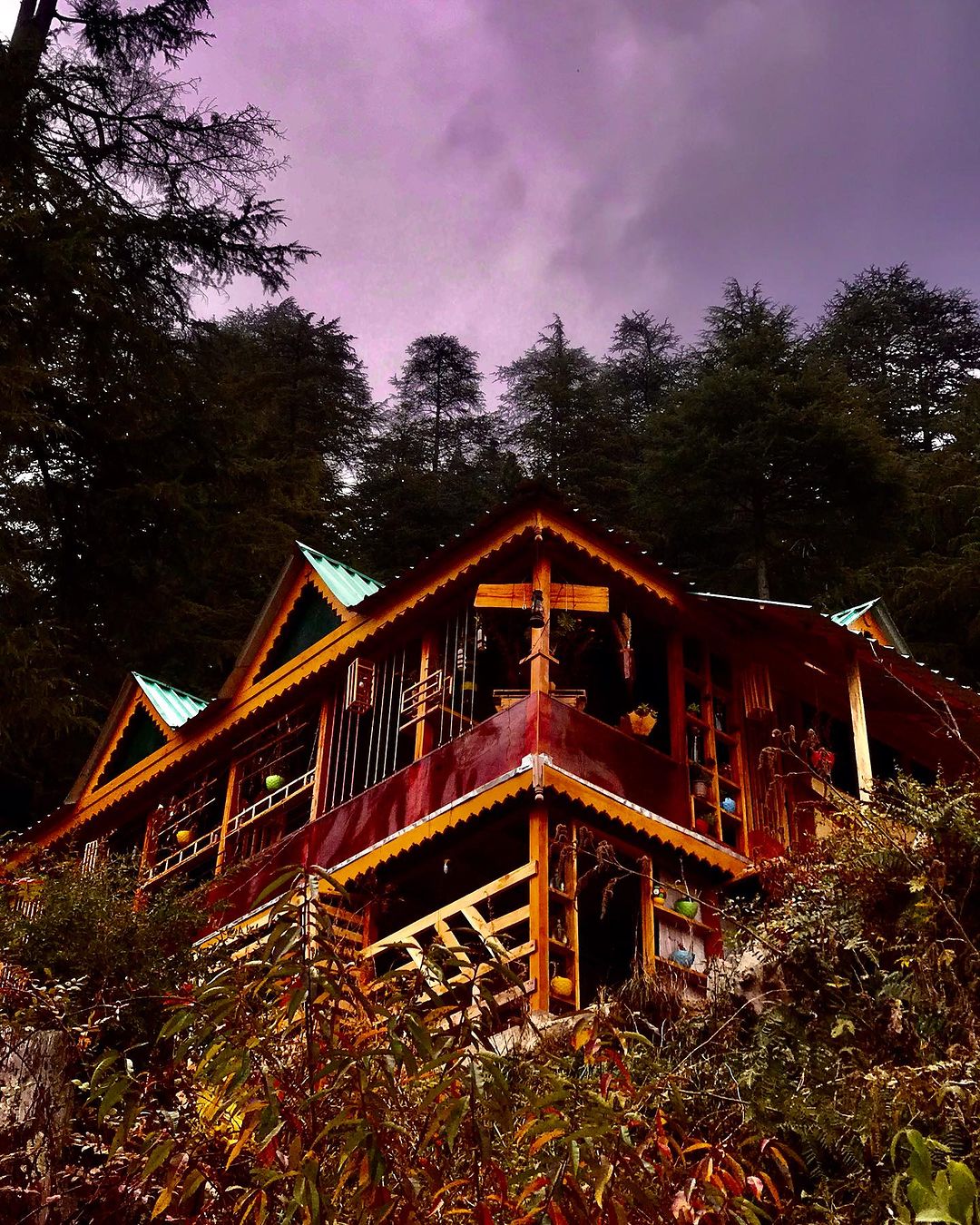 A wooden villa stands against a backdrop of tall trees under a cloudy sky. The structure features a traditional design with a sloped roof, wooden balconies, and bright green accents. The surroundings include lush vegetation, enhancing the natural setting.